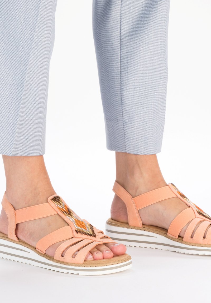 Sandales de couleur pêche avec un design à lanières, présentant un accent en perles et une semelle rayée blanche. Matériau doux avec une finition texturée.