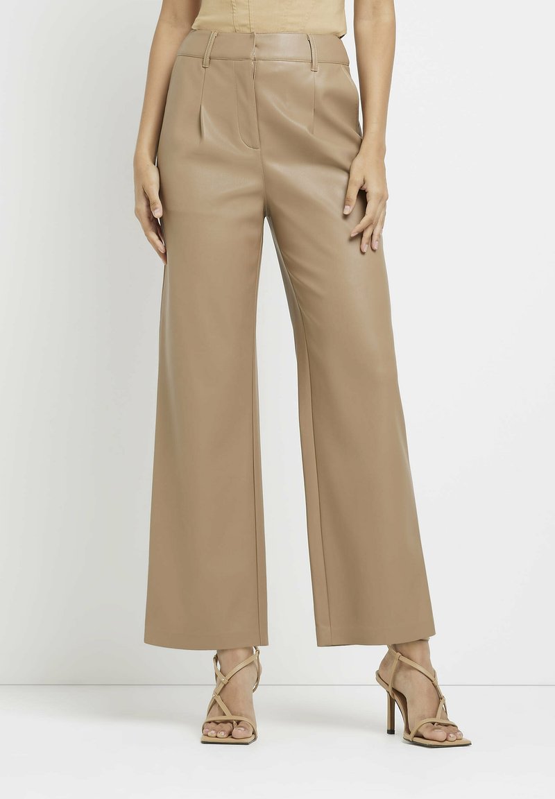 Femme portant un pantalon en cuir beige taille haute à jambes larges, associé à des sandales à talons hauts beiges à brides, debout devant un fond uni.
