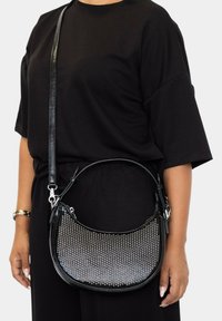 Bolso de hombro de cuero negro con tachuelas plateadas texturizadas, forma curva y correa desmontable. Llevado con un atuendo negro.
