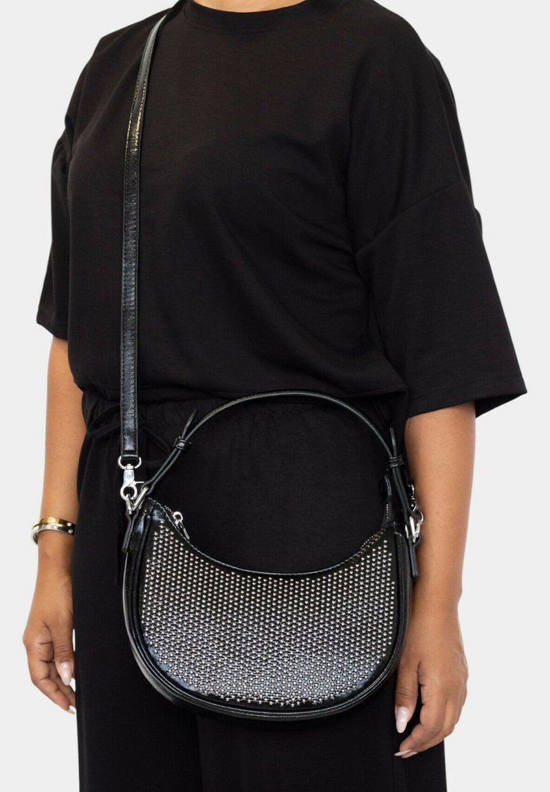 Bolso de hombro de cuero negro con tachuelas plateadas texturizadas, forma curva y correa desmontable. Llevado con un atuendo negro.