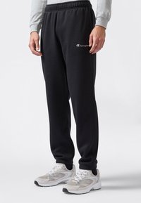 Persona che indossa pantaloni neri Champion e sneakers bianche, in piedi su uno sfondo bianco con le mani rilassate ai lati.