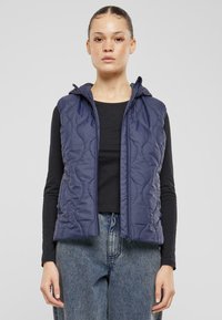 Marinblå vadderad väst med huva, med dragkedja och texturerad tyg. Buren över en svart topp, kombinerad med vida blå jeans.