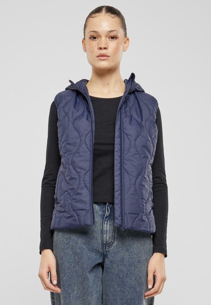 Marinblå vadderad väst med huva, med dragkedja och texturerad tyg. Buren över en svart topp, kombinerad med vida blå jeans.