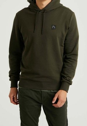 Homme portant un sweat à capuche vert foncé avec un patch logo à l'avant et un pantalon vert foncé assorti, debout sur un fond neutre.