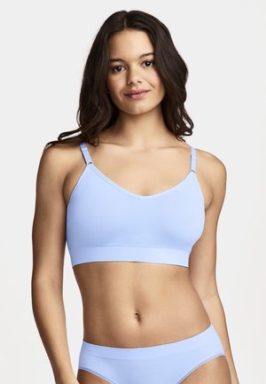 Junge Frau mit langen dunklen Haaren, die einen nahtlosen hellblauen BH und passende Unterwäsche trägt, vor einfachem Hintergrund stehend.