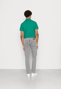 Camiseta polo verde, jeans ajustados grises con un lavado claro y zapatillas blancas, vistas desde atrás. Se aprecian detalles de textura y costura.