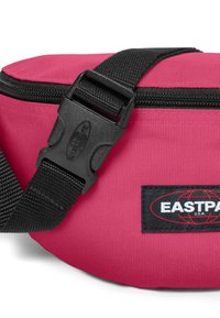 Rosa tygväska med svart dragkedja, justerbar rem och plastspänne. Har en svart logopatch med "EASTPAK"-text. Strukturerad yta.