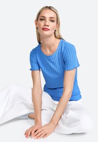 Top azzurro a maniche corte con smock e bordi a scallop, abbinato a pantaloni bianchi a gamba larga.