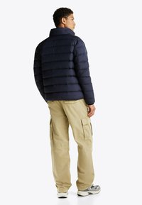 Marineblauwe pufferjack met horizontale stiksels, een opstaande kraag en een gladde textuur. Gecombineerd met beige cargobroek en grijze sneakers.