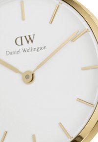 Zlatý hodinky s bielym ciferníkom, ktorý má minimalizované zlaté hodinové značky a ručičky. Názov značky "Daniel Wellington" je zobrazený pod značkou 12.