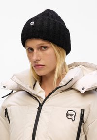 Zwarte gebreide beanie met een ribbelstructuur en een klein wit logo aan de voorkant, gecombineerd met een crèmekleurige puff jas met zwarte accenten.