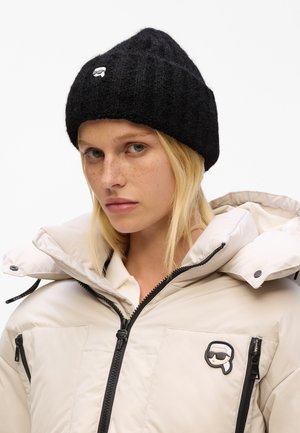 Berretto in maglia nera con una texture a coste e un piccolo logo bianco sul fronte, abbinato a un giubbotto piumino crema con accenti neri.