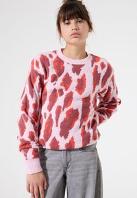 Suéter en rosa, con una textura peluda y un atrevido patrón de estampado animal en rojo y marrón; cuello y puños de canalé. Llevado con jeans grises.
