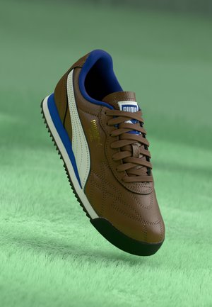 Brauner Puma-Sneaker mit weißem Streifen, blauem Innenfutter und schwarzer Sohle, der über einer grünen, strukturierten Oberfläche schwebt.