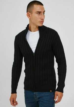 Cárdigan negro con cremallera, que presenta un diseño de canalé, mangas largas, cuello alto y un patrón de punto texturizado. Llevado sobre una camisa blanca y jeans azules.