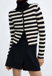 Cardigan rayé noir et crème avec une texture tricotée, encolure ronde et détails en bouton doré, associé à un short noir.