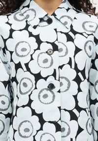 Παλτό με λουλουδάτο σχέδιο που διαθέτει μεγάλα λευκά λουλούδια με γκρι κέντρα σε ανοιχτό μπλε φόντο. Ολοκληρωμένο με γιακά και μαύρα κουμπιά.