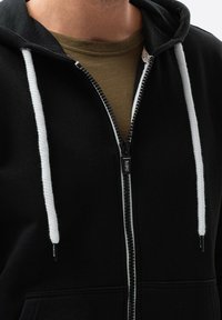 Sweat à capuche noir zippé avec cordons blancs porté par-dessus un T-shirt vert olive à col rond, montrant la partie supérieure du torse et du cou.