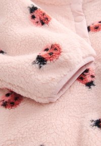 Minkštas rožinis fliso audinys su juodais ir raudonais boružės raštais bei spaudžiamo mygtuko detalė prie apykaklės krašto.