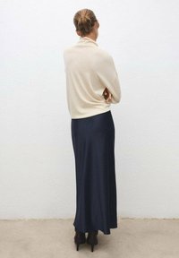 Pull en tricot couleur crème avec une coupe décontractée, associé à une jupe satin bleu marine, longueur sol. Des chaussures à talons hauts marron complètent la tenue.