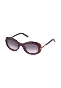 Lunettes de soleil ovales rouge foncé avec des verres gris dégradés et des branches noires avec des accents en or rose sur fond blanc.