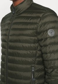 Groene pufferjas met horizontale stiknaden, hoge kraag, ritssluitingen en een cirkelvormige logo-patch op de linkerschouder. Glad, glanzend textuur.