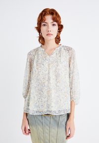 Blusa trasparente di un delicato giallo pallido con motivi floreali blu e arancioni, caratterizzata da un scollo a V e maniche arricciate, abbinata a una gonna di jeans.