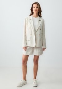 Leinenmischung Blazer in hellbeige mit doppelter Knopfleiste, passenden Shorts und weißen Sneakers. Glatte Textur, lockere Passform, minimale Details.