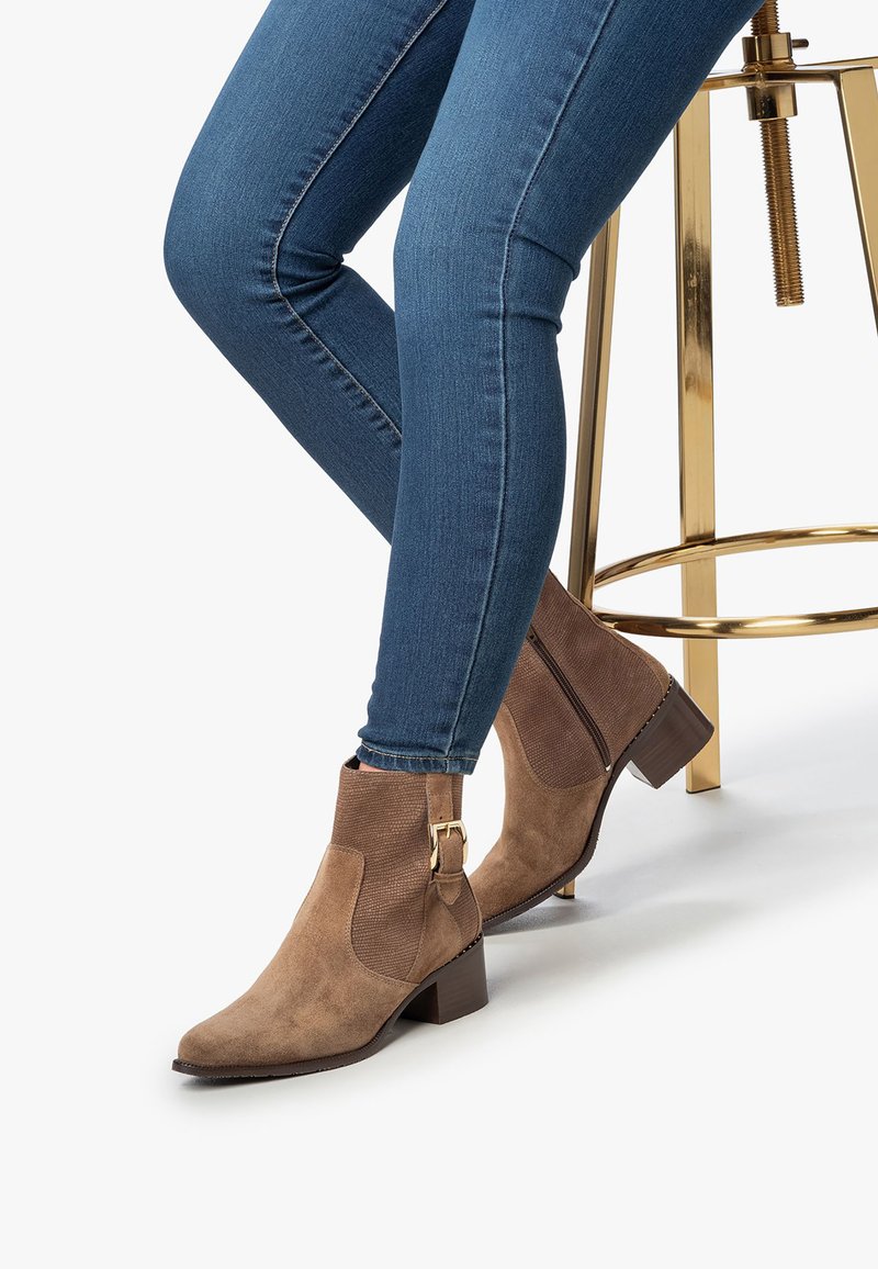Piernas en jeans ajustados azules y botas de ante marrón con tacones cuadrados, sentadas en un taburete de metal dorado brillante.