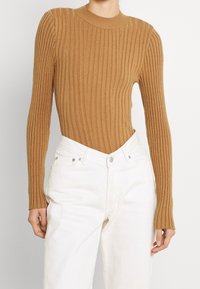 Braun geripptes Strickoberteil mit langen Ärmeln und figurbetontem Schnitt. Kombiniert mit hoch taillierten weißen Hosen mit Fünf-Taschen-Design und silbernen Beschlägen.