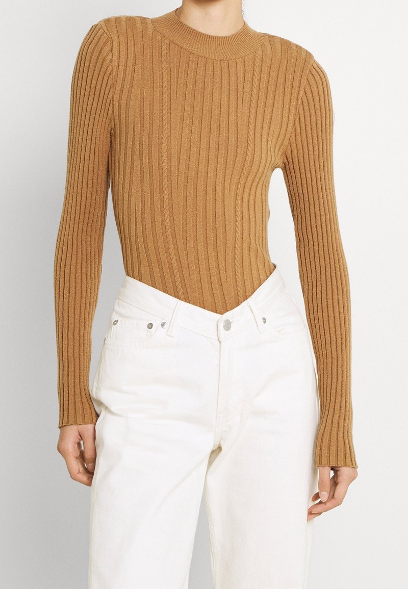 Braun geripptes Strickoberteil mit langen Ärmeln und figurbetontem Schnitt. Kombiniert mit hoch taillierten weißen Hosen mit Fünf-Taschen-Design und silbernen Beschlägen.