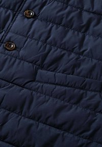 Marineblaue Steppweste mit gesteppter Textur, ausgestattet mit zwei braunen Knöpfen und einer versteckten Tasche. Das Material wirkt weich und isoliert.