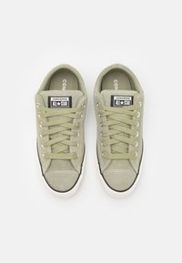 Zapatillas de lona caqui con punta redonda, suela de goma blanca, costuras grises y parche de logo negro. Presentan cordones planos y un diseño de corte bajo.