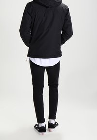 Černá kapucňová bunda s hladkou texturou, nošená přes bílou košili, kombinovaná s černými úzkými džíny a černými botami s bílými akcenty.