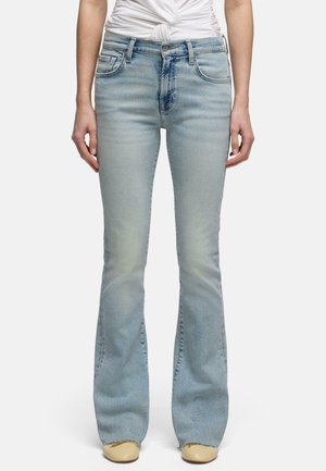 Frau trägt hellblaue hoch taillierte Schlagjeans, off-white geknotetes Top und beige Schuhe, steht vor einem schlichten Hintergrund.