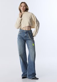 Femme portant un pull en maille torsadée crème court, un jean taille haute à jambes larges avec un accessoire de chaîne, et des chaussures noires à talons pointus sur un fond blanc.