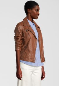 Veste de motard en cuir marron avec des poches zippées à l'avant, des épaulettes et une silhouette ajustée, portée sur une chemise rayée bleu et blanc.