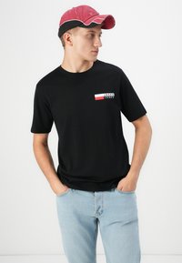 Zwarte T-shirt met korte mouwen, featuring een wit en rood logo detail op de linkerkant van de borst. Draagt het met lichtblauwe jeans en een rode pet.