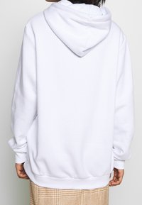 Vit oversized hoodie med snörning i huood, ribbade ärmar och nederkant. Mjuk, slät textur; bakvy visar inga ytterligare designelement.