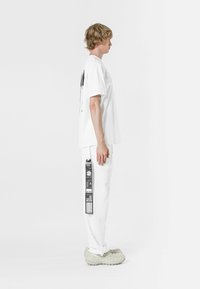 Camiseta blanca de gran tamaño con un gráfico negro en la parte posterior, combinada con pantalones blancos con impresiones laterales negras, llevada con zapatos beige texturizados.