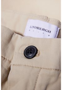 Bouton noir cousu sur un tissu beige près d'une étiquette de vêtement blanche avec le texte « LINDBERGH ».
