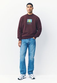 Vinröd sweatshirt med en Keith Haring-grafik; rund halsringning, ribbade muddar och fåll. Kombinerad med ljusblå denimjeans och vita sneakers.