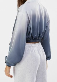 Giacca accorciata blu ombre in tessuto morbido, vita elastica, maniche arricciate e colletto classico. Indossata sopra pantaloni della tuta grigi.