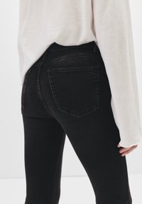 Jean en denim noir à coupe slim, avec une taille haute, deux poches arrière et un détail de couture minimal, associé à un haut de couleur claire.