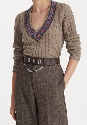 Personne portant un pull beige en maille torsadée avec un col en V à motif et un pantalon en tweed marron avec une ceinture foncée et un accessoire chaîne argentée.
