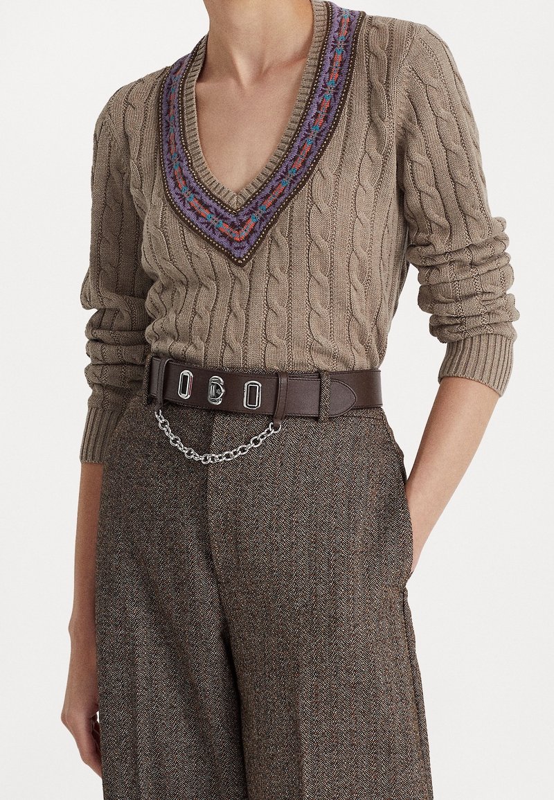 Personne portant un pull beige en maille torsadée avec un col en V à motif et un pantalon en tweed marron avec une ceinture foncée et un accessoire chaîne argentée.