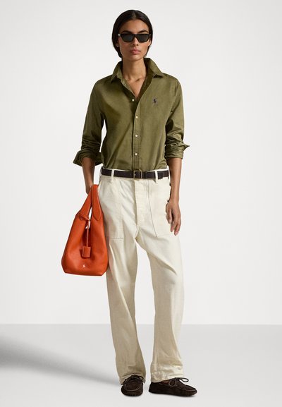 Nő, aki olívazöld gombos inget, krémszínű, széles szárú nadrágot, sötétbarna övet és mokaszint visel, narancssárga bőr kézitáskát hord, valamint fekete napszemüveget.