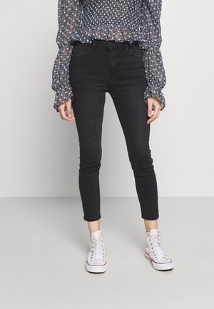 Personne portant un jean skinny noir, des baskets blanches montantes et un chemisier noir transparent à pois blancs, debout devant un fond uni.