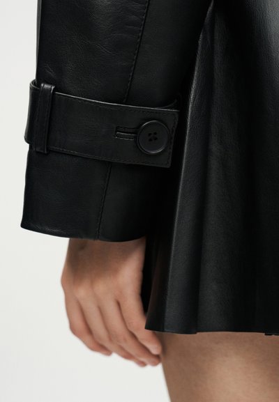 Μανίκι μαύρου δερμάτινου παλτού με κουμπωμένο λουράκι, που καλύπτει μερικώς ένα χέρι και μηρό πάνω σε ένα απλό ανοιχτό φόντο.