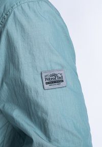 Jachetă bleu deschis, cu un material texturat, având un abțibild rectangular cu inscripția "1989 Petrol Ind" și detalii suplimentare.
